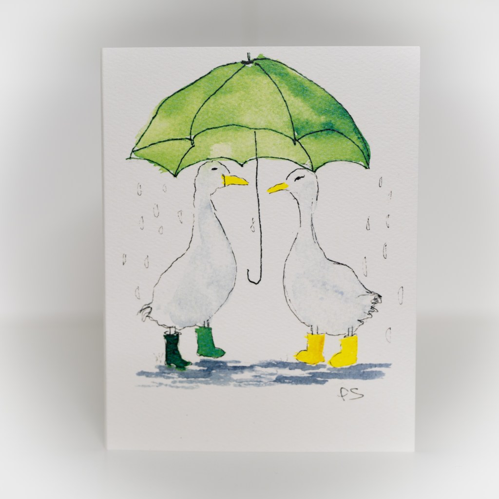 A greeting card featuring a painting of two ducks wearing boots under an umbrella in the rain.